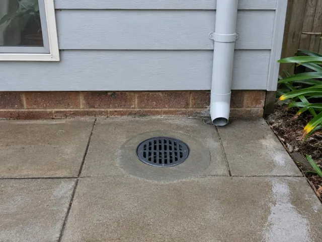 Outdoor drain grate set in dry paved patio beside grey weatherboard wall in Eastlakes